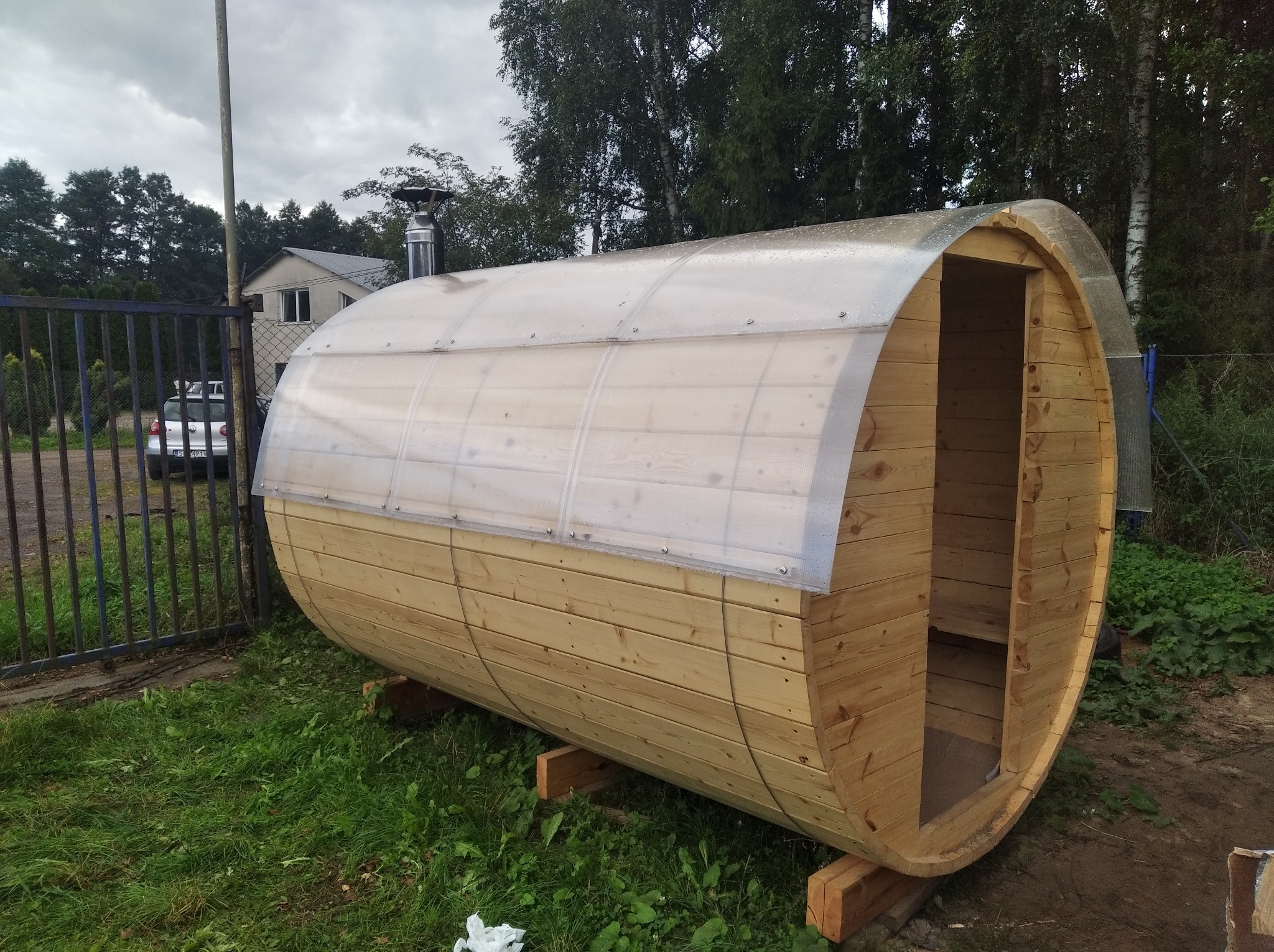 Drewniana sauna w kształcie beczki z przezroczystym dachem, ustawiona na trawie obok ogrodzenia. Widoczny komin i fragment domu w tle. Nietypowy kształt sauny.