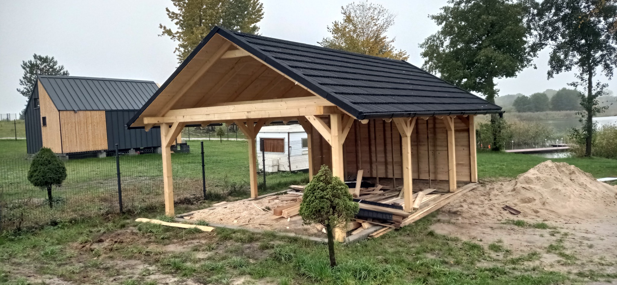 Wznoszona drewniana altana z czarnym dachem na działce, widoczne elementy konstrukcyjne i resztki drewna. W tle dom z bali i jezioro.