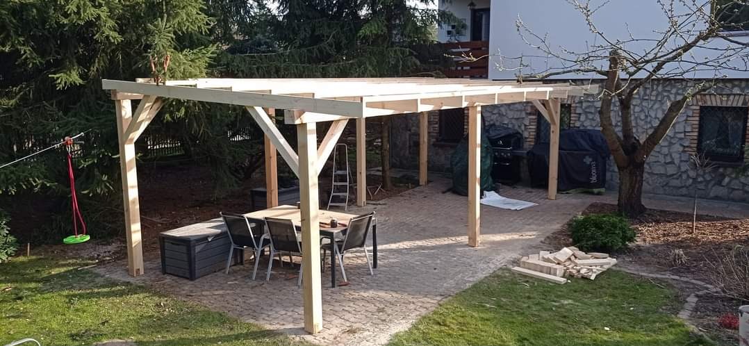 Surowa drewniana pergola w ogrodzie z meblami ogrodowymi i grillem w tle. Widoczna huśtawka na linie. Konstrukcja w trakcie montażu, z elementami do wykończenia.