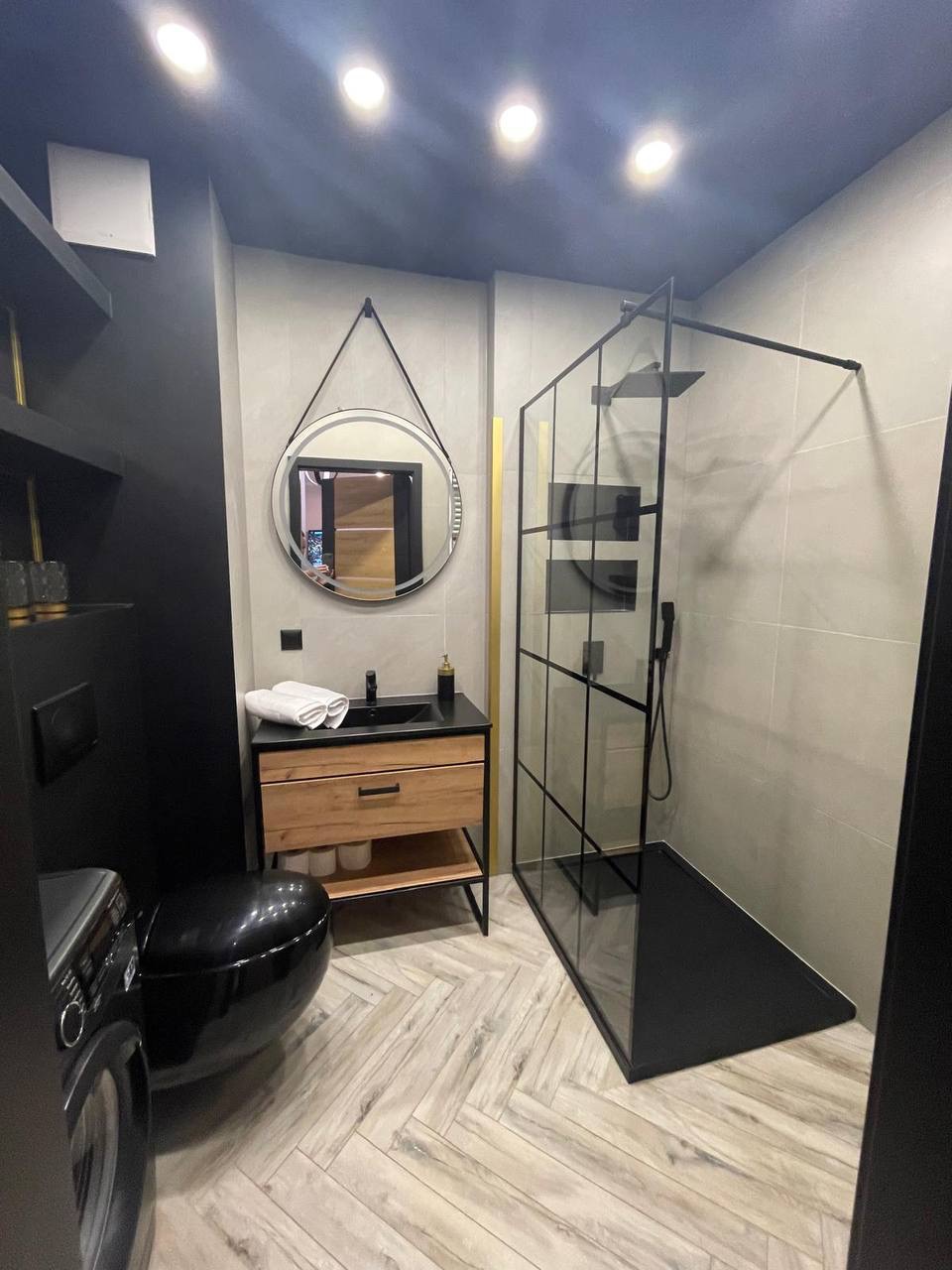 Nowoczesna łazienka z kabiną typu walk-in, drewnianą szafką pod umywalką i okrągłym lustrem na tle płytek w odcieniach szarości i beżu. Uwagę przykuwa czarna armatura.