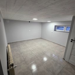 Usługi Remontowo Budowlane Maciej Burz - Wnętrze po remoncie: szare płytki podłogowe, świeżo pomalowane szare ściany, surowy betonowy sufit z oświetleniem punktowym i okno piwniczne z kratą.
