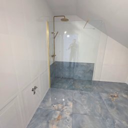 Usługi Remontowo Budowlane Maciej Burz - Nowoczesna kabina prysznicowa ze złotymi elementami w łazience na poddaszu, wykończona płytkami imitującymi marmur. Widoczne detale armatury i szklana ścianka.