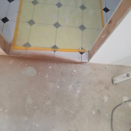 Usługi Remontowo Budowlane Maciej Burz - Fragment podłogi z częściowo ułożonymi płytkami w czarno-białą szachownicę, oddzielonymi żółtą taśmą od surowego betonu. Widoczne buty i poziomica.