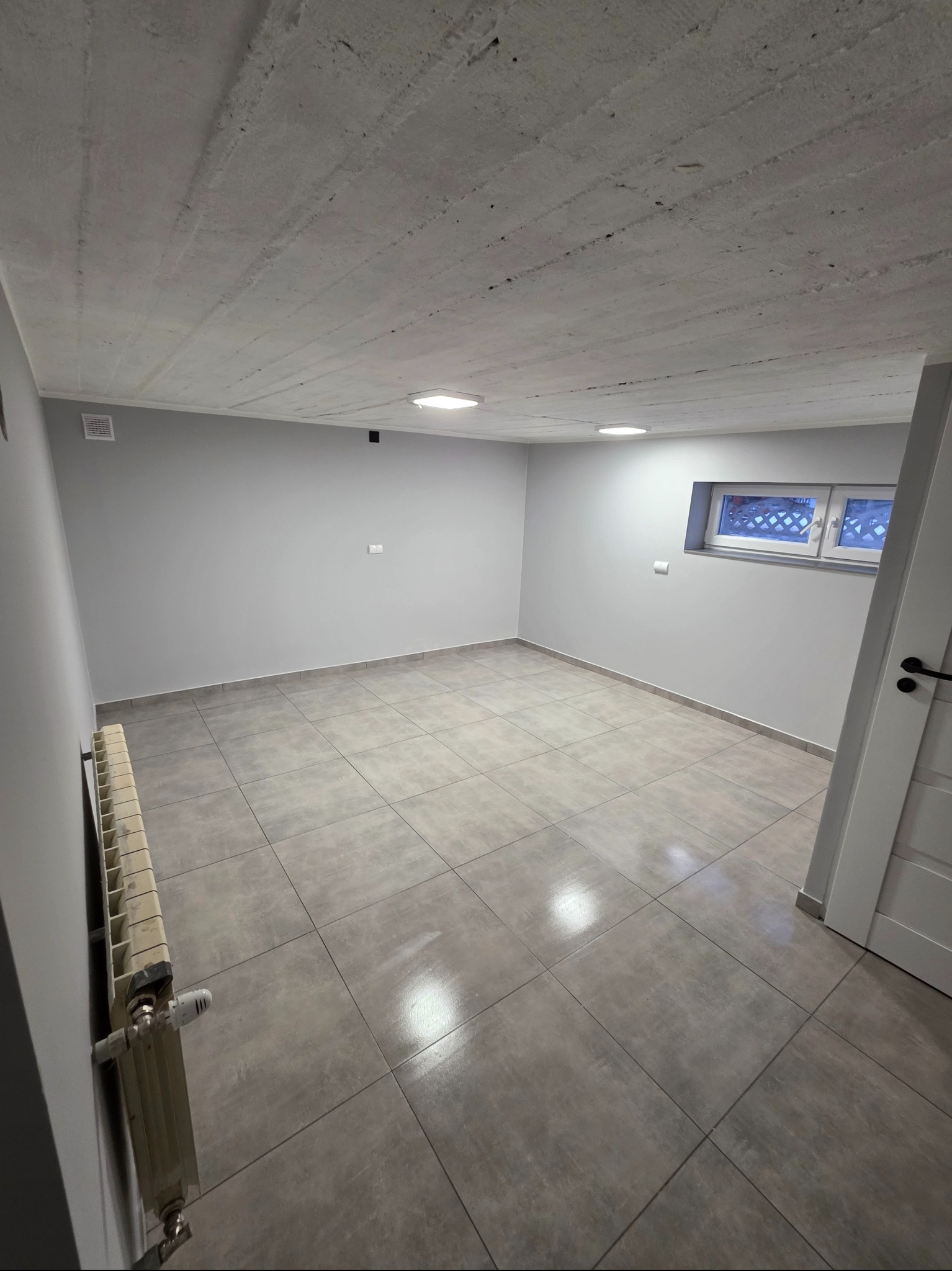 Wnętrze po remoncie: szare płytki podłogowe, świeżo pomalowane szare ściany, surowy betonowy sufit z oświetleniem punktowym i okno piwniczne z kratą.