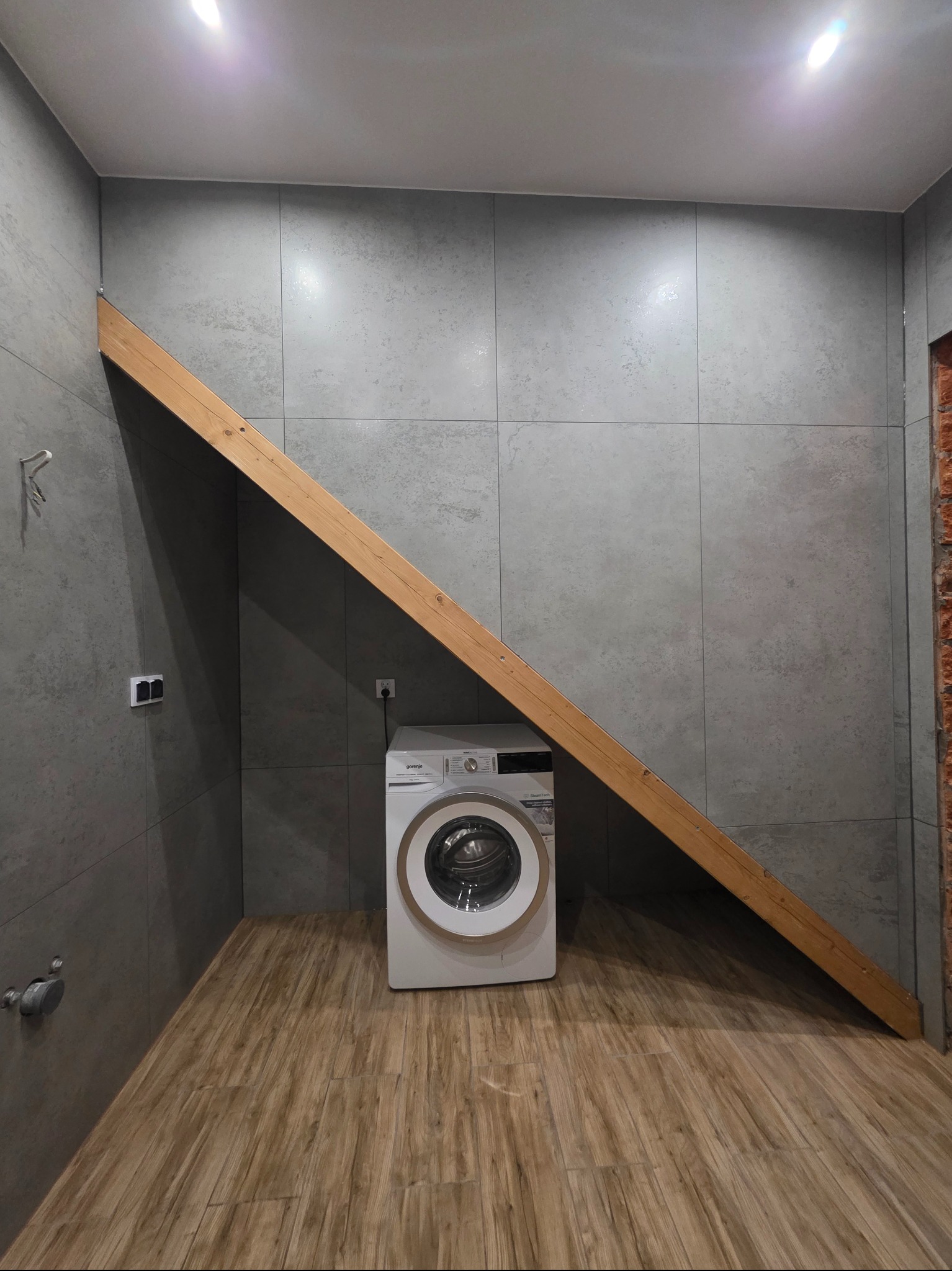 Wykończona łazienka z szarą płytką, drewnianą podłogą i białą pralką. Skośna belka pod sufitem dodaje unikalnego charakteru wnętrzu. Widoczne gniazdka i fragment ściany z cegły.