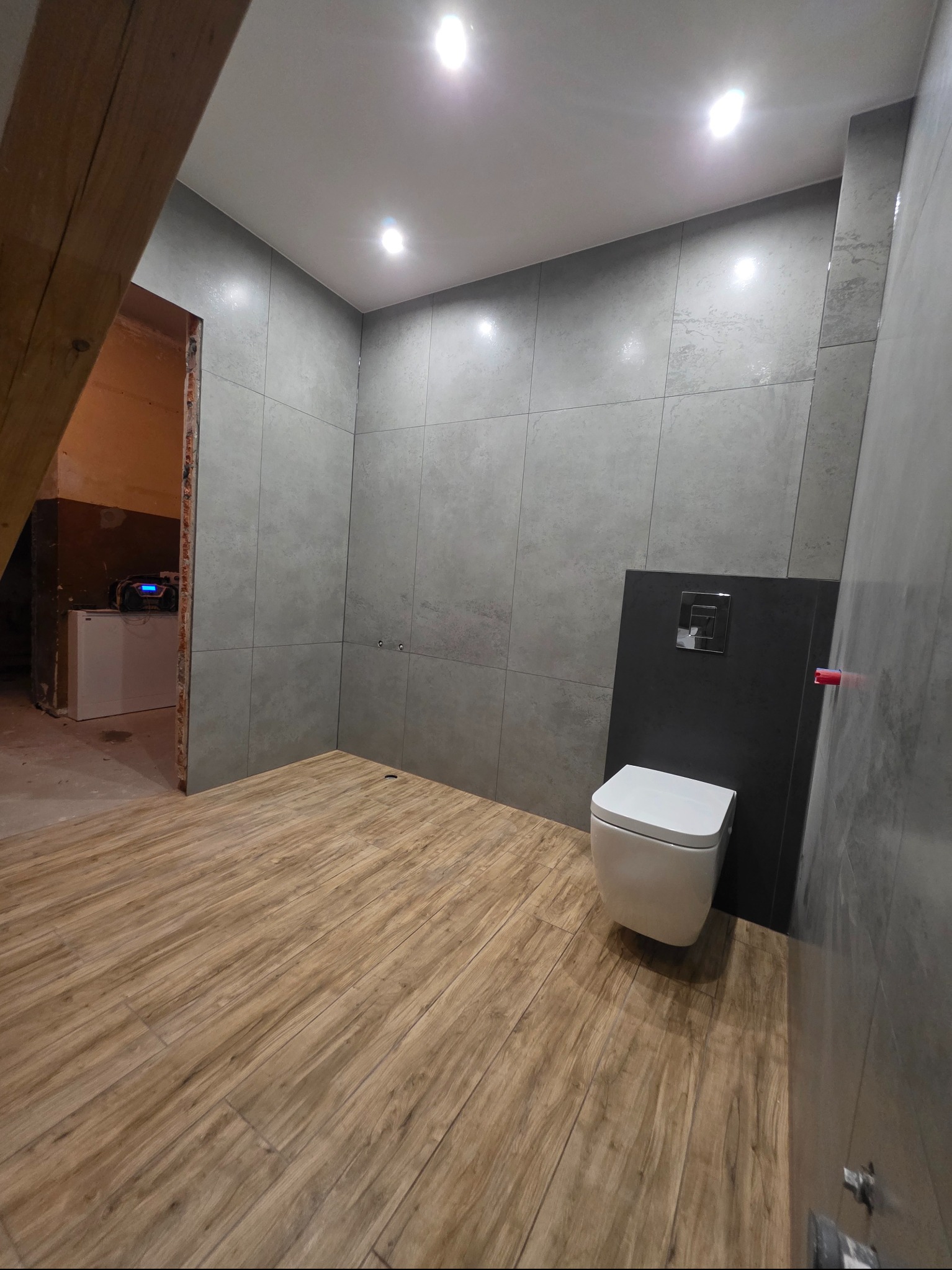 Nowoczesna łazienka w trakcie remontu: szare płytki ścienne, drewnopodobna podłoga, podwieszana toaleta. Widoczna belka stropowa i sprzęt w sąsiednim pomieszczeniu.
