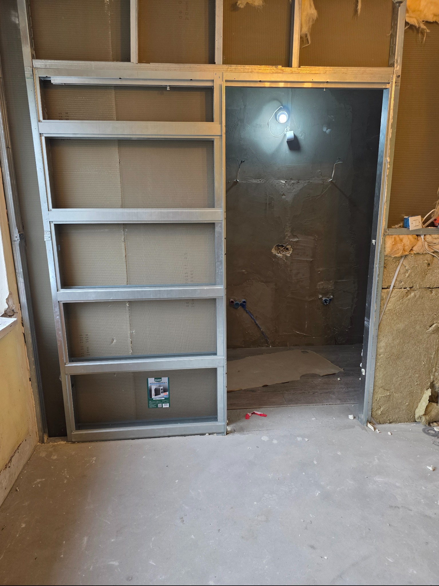 Konstrukcja metalowa z płytami gipsowymi w trakcie montażu, widoczne instalacje w ścianie. Prace wykończeniowe w łazience, surowy stan.