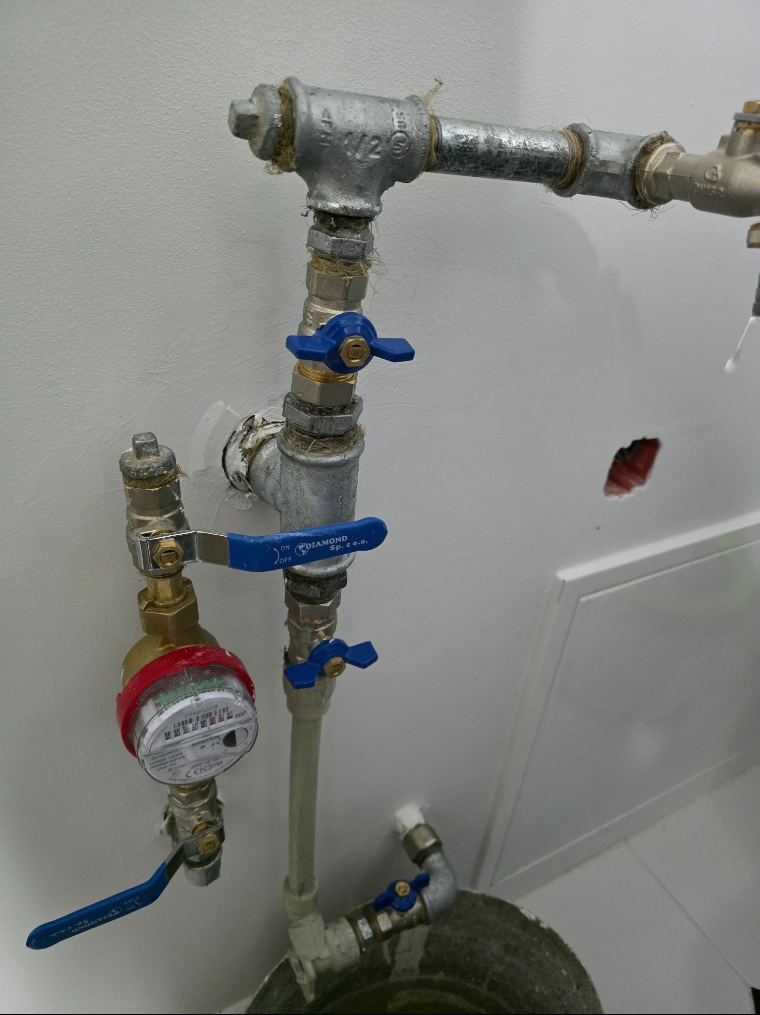 Instalacja wodna z licznikiem i zaworami odcinającymi na tle białej ściany. Widoczne ślady po naprawach i niedokładności w wykończeniu.