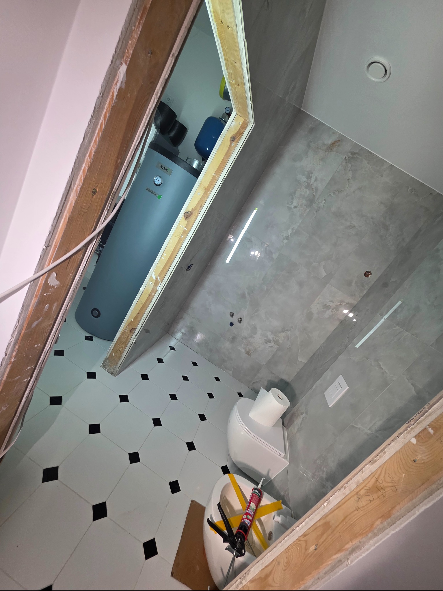 Wnętrze łazienki w trakcie remontu: widoczny bojler, biała toaleta z rolką papieru, szare płytki na ścianach i biała podłoga z czarnymi akcentami. Surowa rama drzwi.