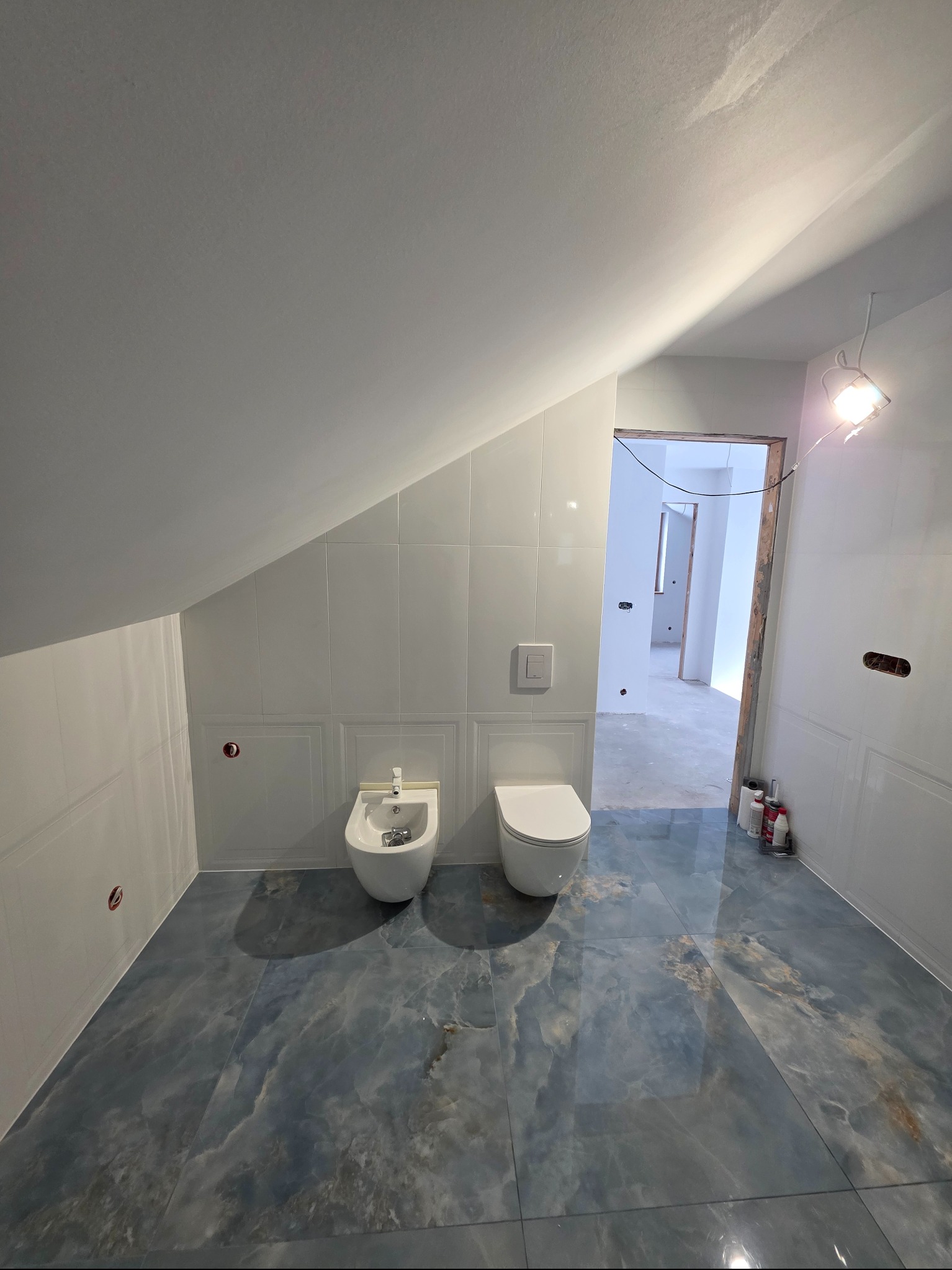 Biała łazienka na poddaszu z bidetem i toaletą, wykończona płytkami imitującymi marmur. Widoczne przygotowane instalacje elektryczne i oświetlenie robocze.