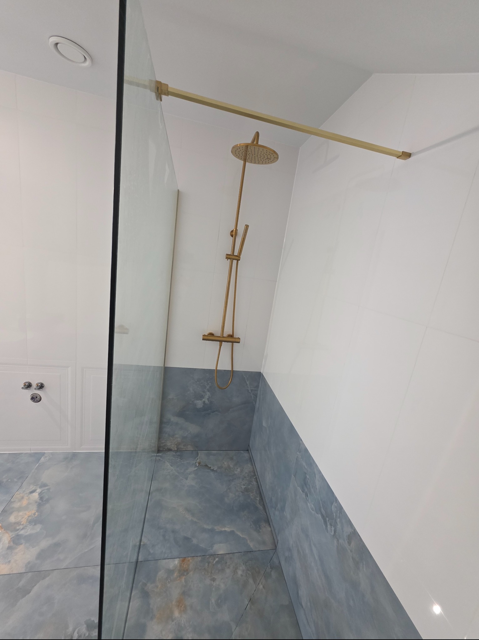 Nowoczesna kabina prysznicowa z złotą armaturą i szklaną ścianką, wykończona białymi i niebieskimi płytkami imitującymi marmur. Minimalistyczny design.