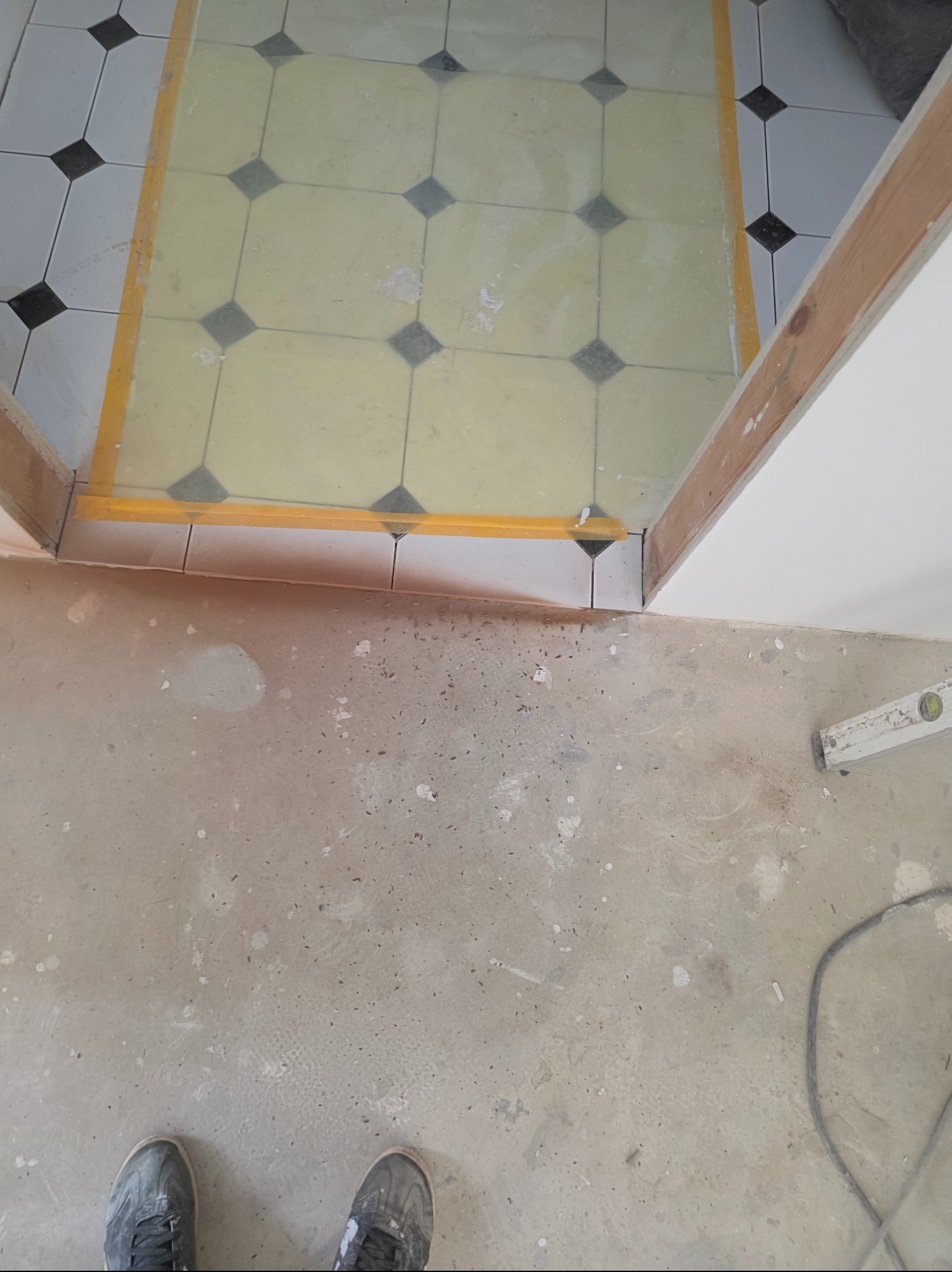 Fragment podłogi z częściowo ułożonymi płytkami w czarno-białą szachownicę, oddzielonymi żółtą taśmą od surowego betonu. Widoczne buty i poziomica.