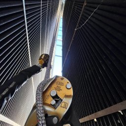 Alpine RS ROBERT SŁABAK - Praca wysokościowa przy elewacji budynku z użyciem sprzętu Petzl RIG. Widok z dołu na linę i urządzenie asekuracyjne na tle żaluzji fasadowych.