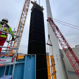 Alpine RS ROBERT SŁABAK - Montaż elementu konstrukcyjnego na wysokości z użyciem dwóch dźwigów. Pracownik w kasku i odblaskowej kamizelce nadzoruje z rusztowania. W tle komin i pochmurne niebo.
