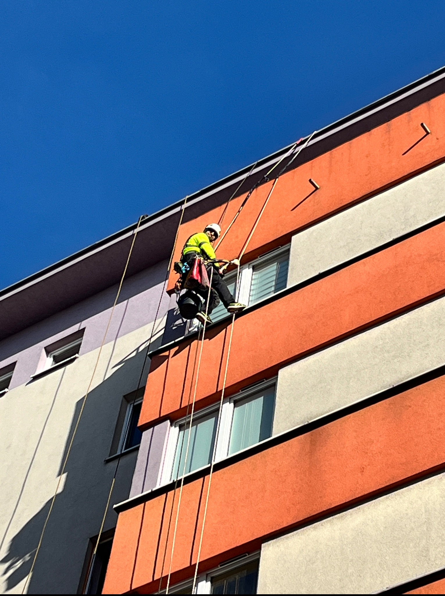 Pracownik w uprzęży na linie, w kasku i jaskrawej kurtce, na tle pomarańczowo-szarej elewacji budynku podczas dnia słonecznego. Ujęcie z dołu.