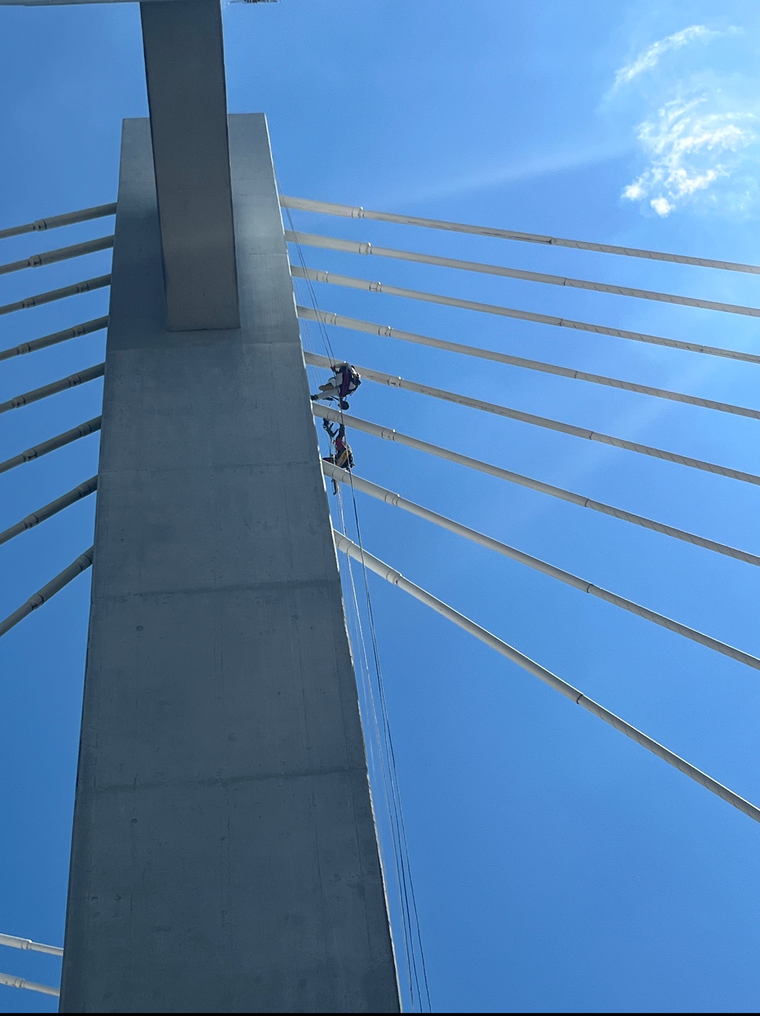Pracownicy na linach zabezpieczających, wykonujący prace na wysokościach na betonowym pylonie mostu w słoneczny dzień. Widok z dołu, niebo w tle.
