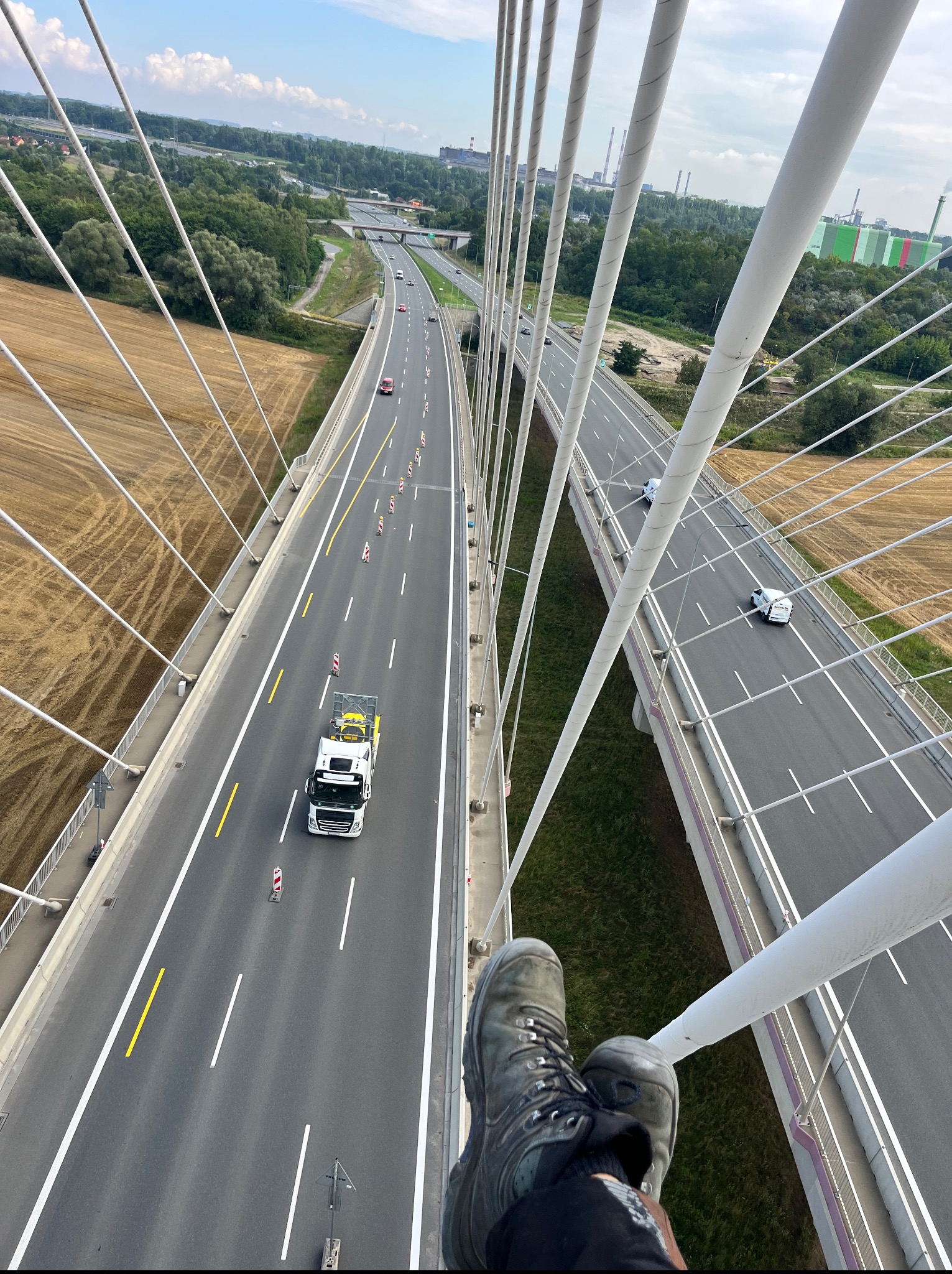 Widok z góry na autostradę z perspektywy osoby stojącej na konstrukcji mostu linowego. W tle widać pola, lasy i zabudowania przemysłowe.