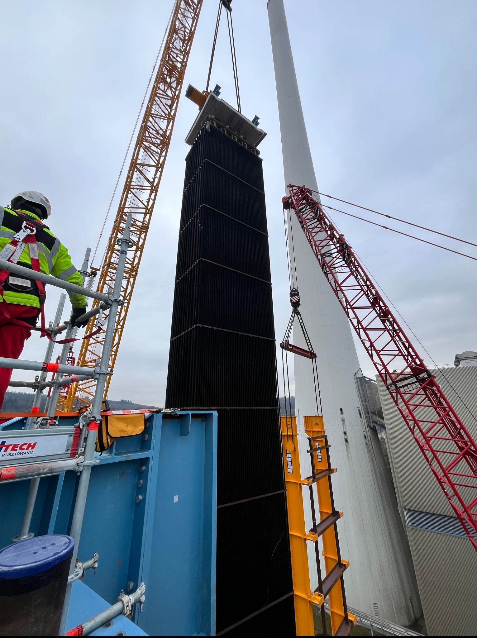 Montaż elementu konstrukcyjnego na wysokości z użyciem dwóch dźwigów. Pracownik w kasku i odblaskowej kamizelce nadzoruje z rusztowania. W tle komin i pochmurne niebo.