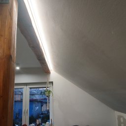 Greenvolt Service Franciszek Jaworski - Wnętrze poddasza z drewnianą belką i oświetleniem LED, widok na okno z rośliną wiszącą. Dwustrefowe oświetlenie z efektem rozjaśniania.