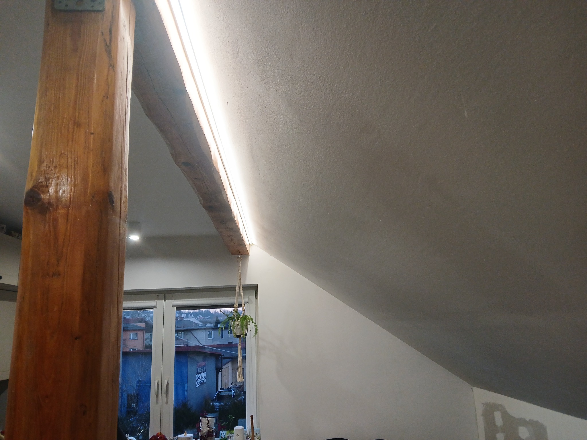 Wnętrze poddasza z drewnianą belką i oświetleniem LED, widok na okno z rośliną wiszącą. Dwustrefowe oświetlenie z efektem rozjaśniania.