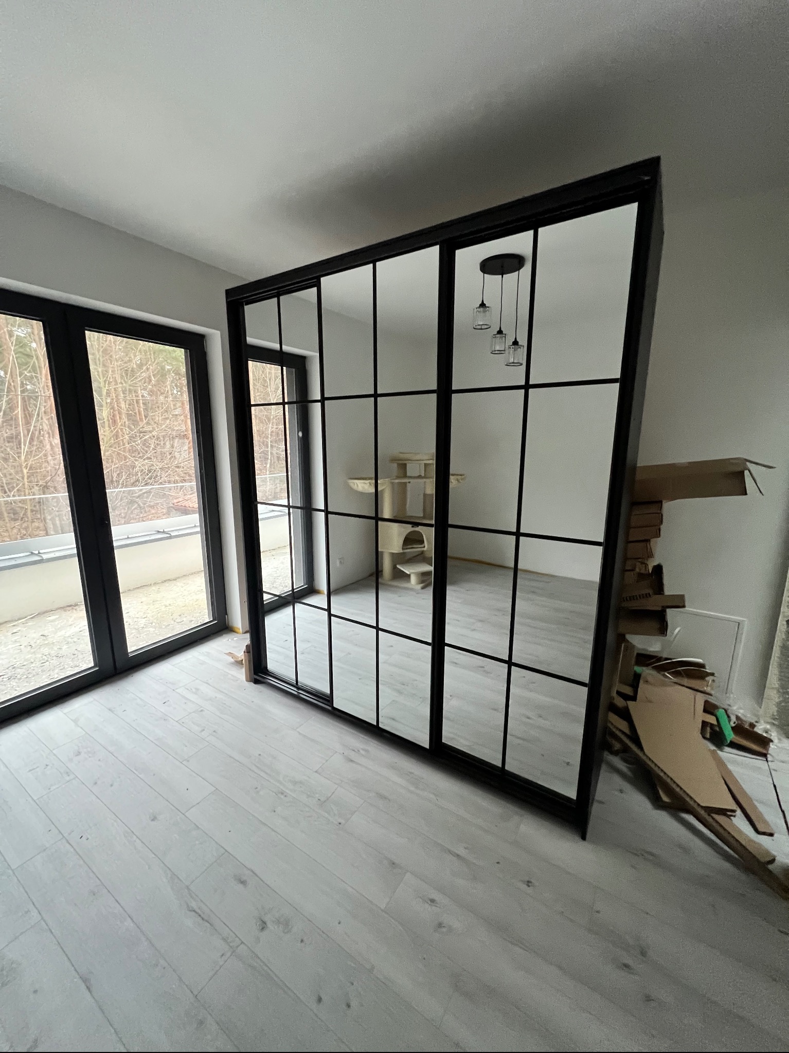 Nowoczesna szafa przesuwna z czarnymi ramami i lustrzanymi drzwiami, widoczna w jasnym pokoju z panelami podłogowymi. W tle okno i elementy wyposażenia.