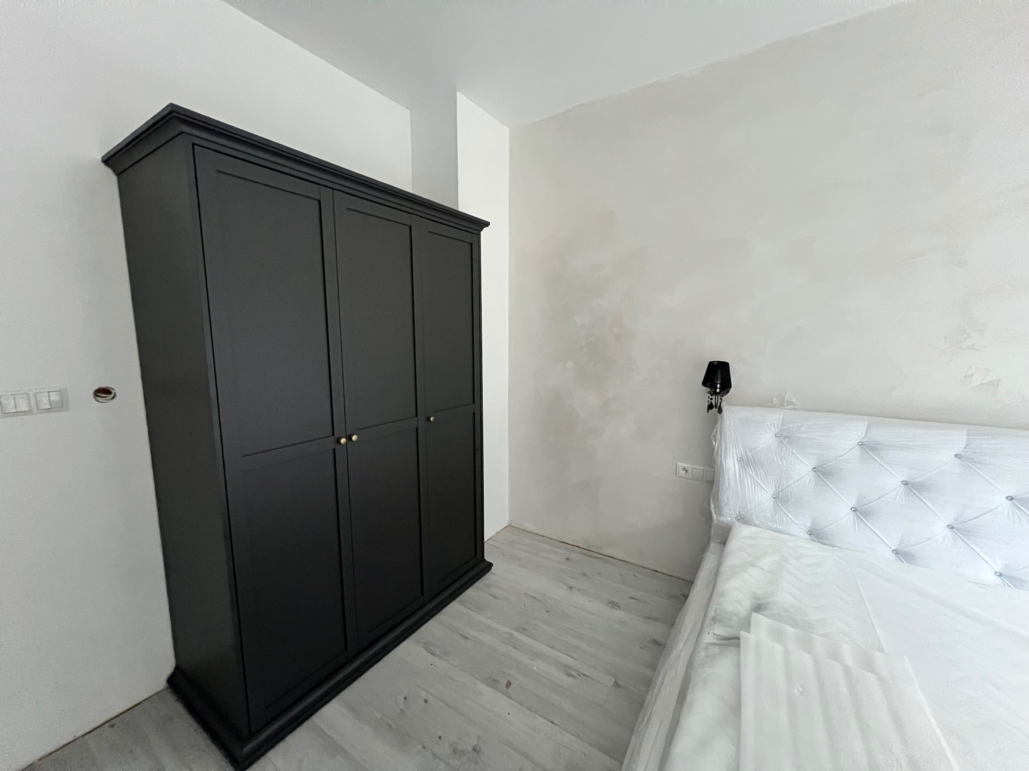 Czarna, trzydrzwiowa szafa z dekoracyjnymi frezami i złotymi uchwytami, ustawiona w rogu pokoju z jasną podłogą i łóżkiem z zagłówkiem w folii ochronnej.