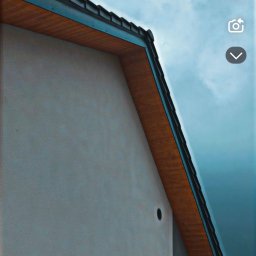 Z.U.H Jakub Sitarski - Elewacja budynku z drewnianym podbitką dachu na tle błękitnego nieba. Widoczny fragment okna z żaluzjami i otwór wentylacyjny.