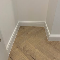 Z.U.H Jakub Sitarski - Ukończone wykończenie podłogi z wzorem jodełki, listwy przypodłogowe w narożniku, kremowe ściany. Detal wykończenia wnętrza.