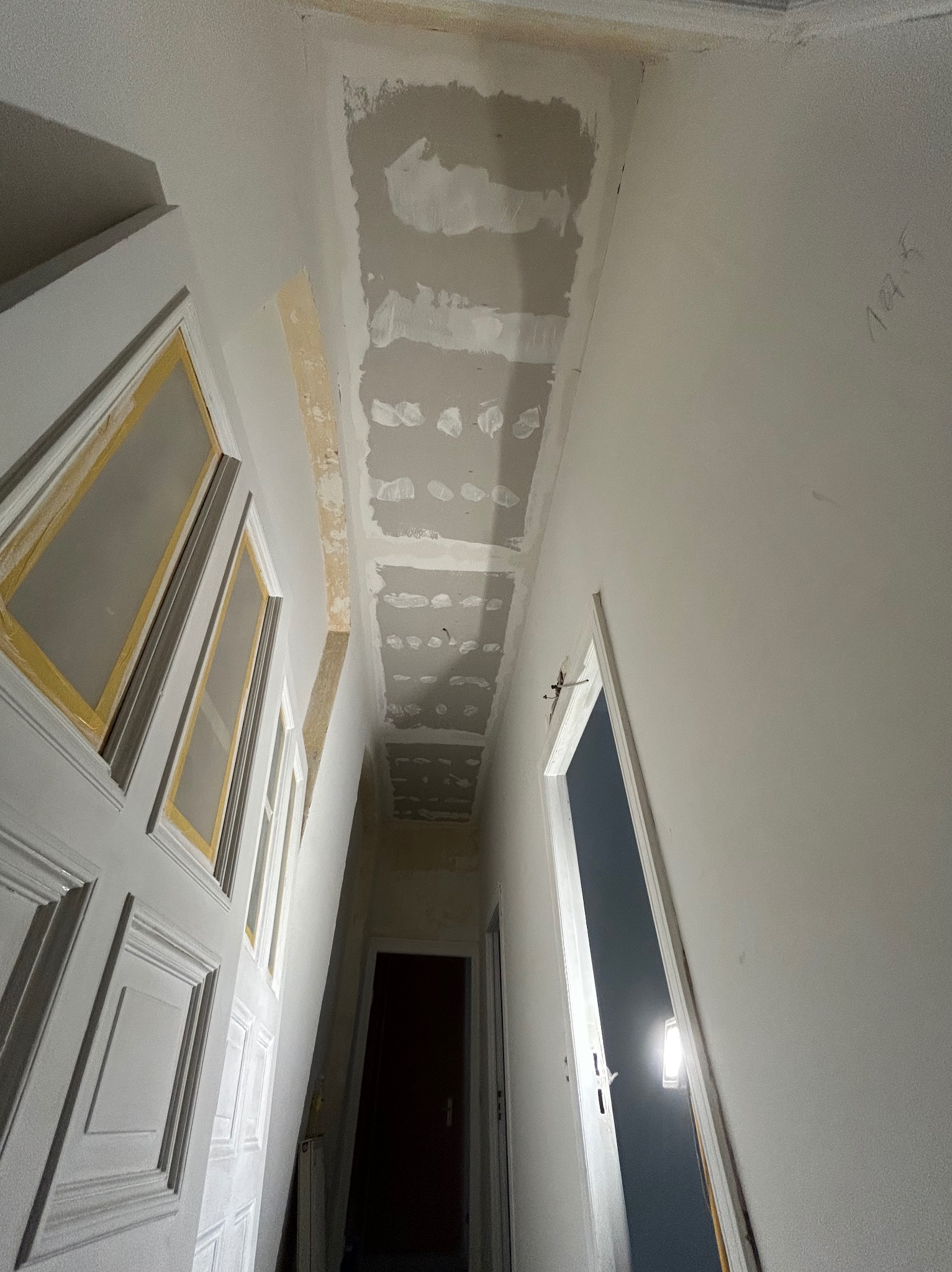 Korytarz w trakcie prac. Widoczny sufit po szpachlowaniu, przygotowany do dalszych prac wykończeniowych. Drzwi zabezpieczone taśmą malarską.
