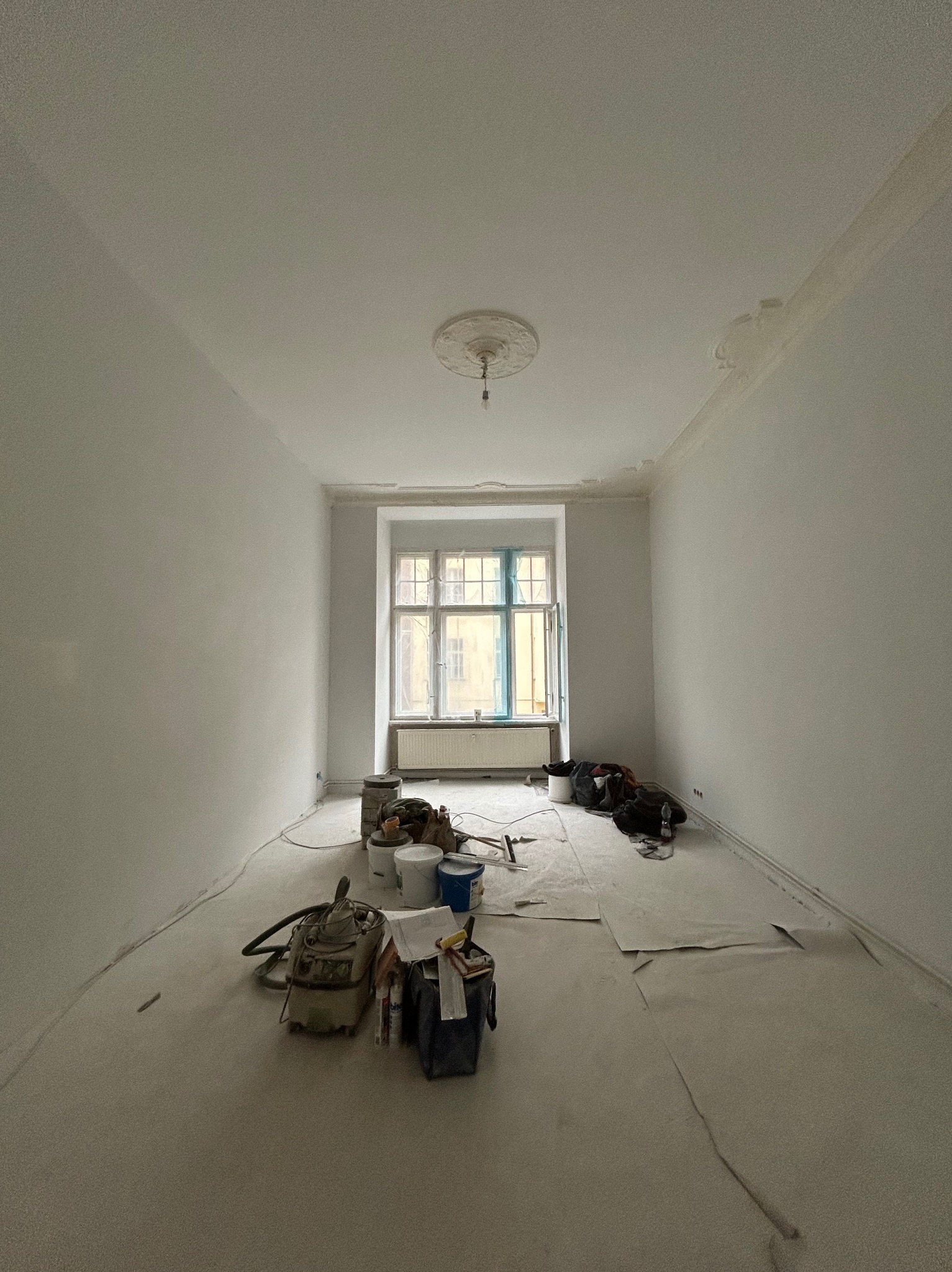 Jasne wnętrze pokoju po malowaniu ścian na biało, widoczne narzędzia malarskie i zabezpieczenia podłogi. Okno z niebieską folią ochronną.