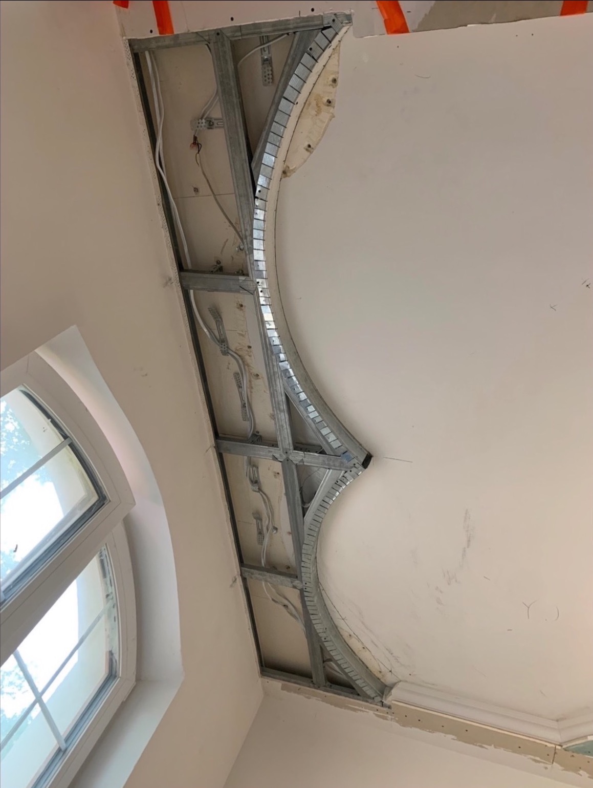 Kreatywna konstrukcja sufitu podwieszanego z metalowym stelażem i instalacją elektryczną, widoczna z perspektywy żabiej, w jasnym wnętrzu z oknem.