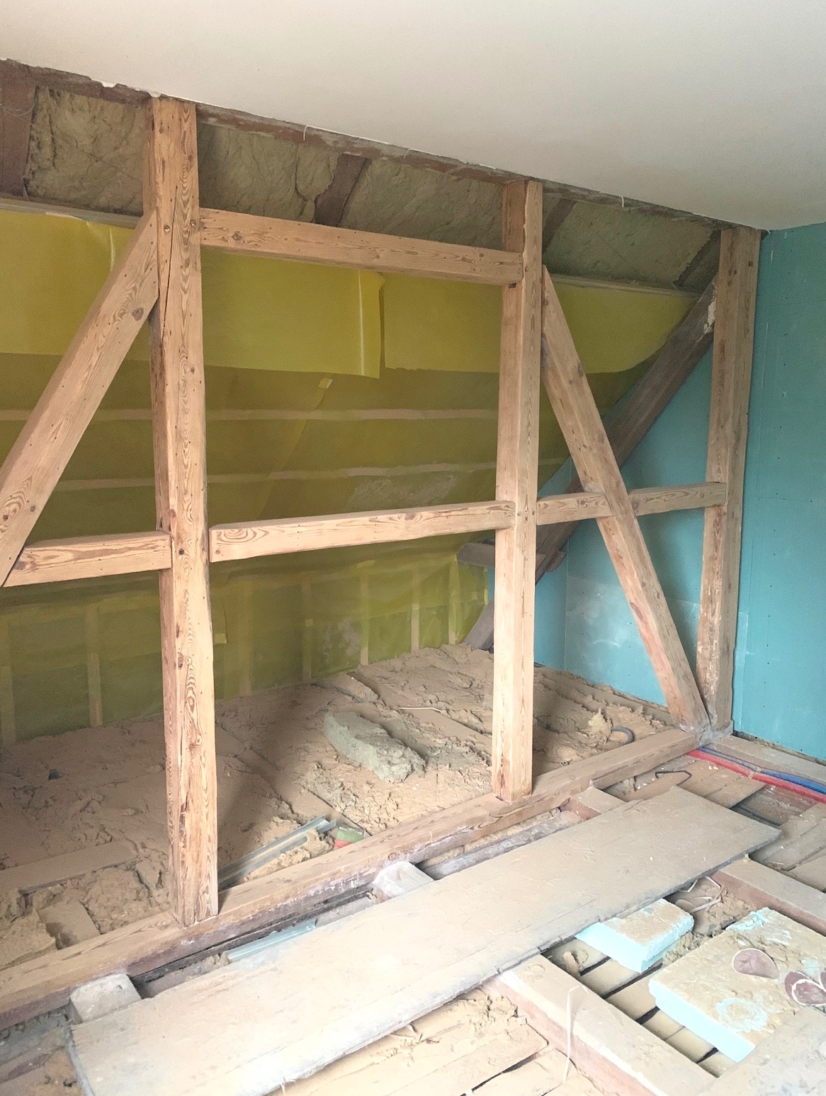Drewniana konstrukcja szkieletowa ściany z żółtą izolacją w tle, widoczne fragmenty podłogi i instalacji. Etap prac wykończeniowych w pomieszczeniu z turkusową ścianą.