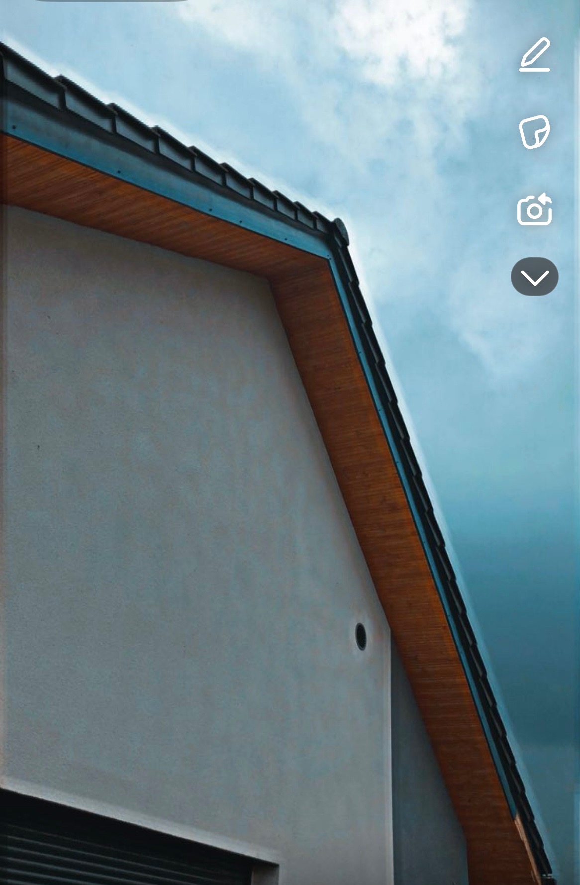 Elewacja budynku z drewnianym podbitką dachu na tle błękitnego nieba. Widoczny fragment okna z żaluzjami i otwór wentylacyjny.