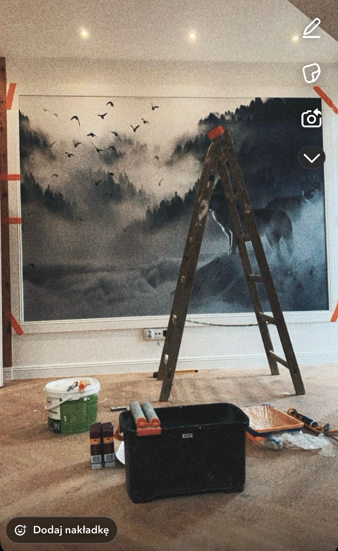 Malowanie ściany z motywem leśnym. Widoczna drabina, kuweta malarska i wałki. Prace wykończeniowe w pomieszczeniu z beżową podłogą i białymi listwami przypodłogowymi.