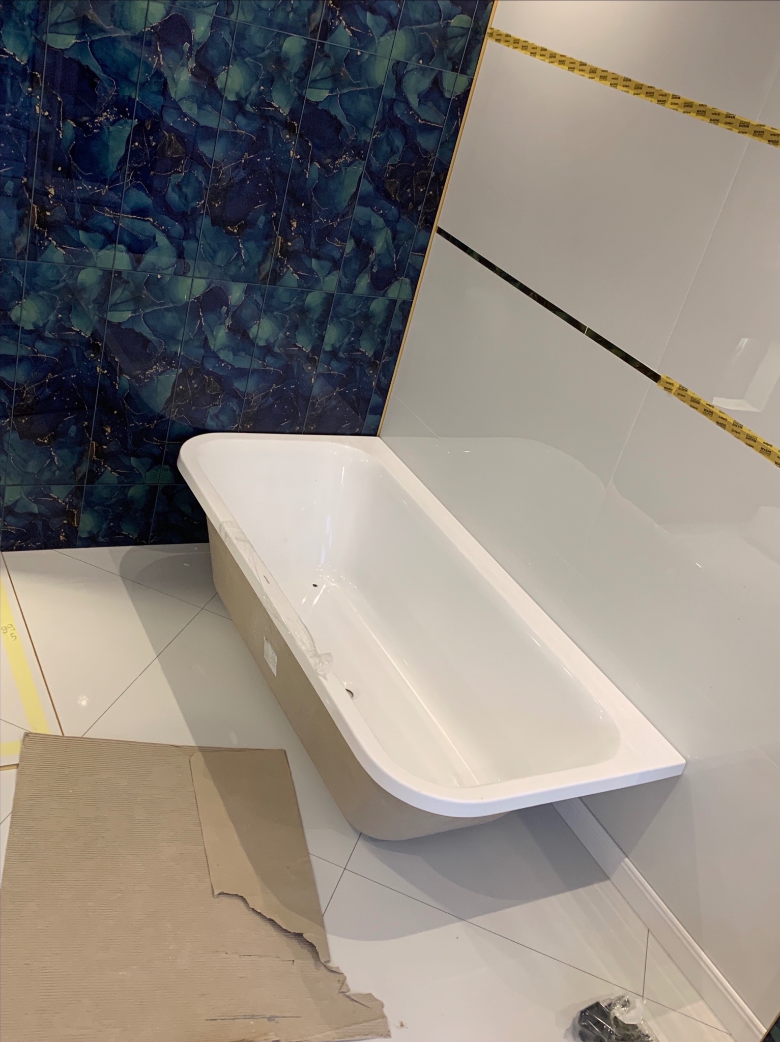 Biała wanna w trakcie montażu w łazience z dekoracyjnymi, ciemnymi płytkami imitującymi marmur i białymi płytkami z czarnym akcentem. Widoczne narzędzia i fragmenty materiałów.