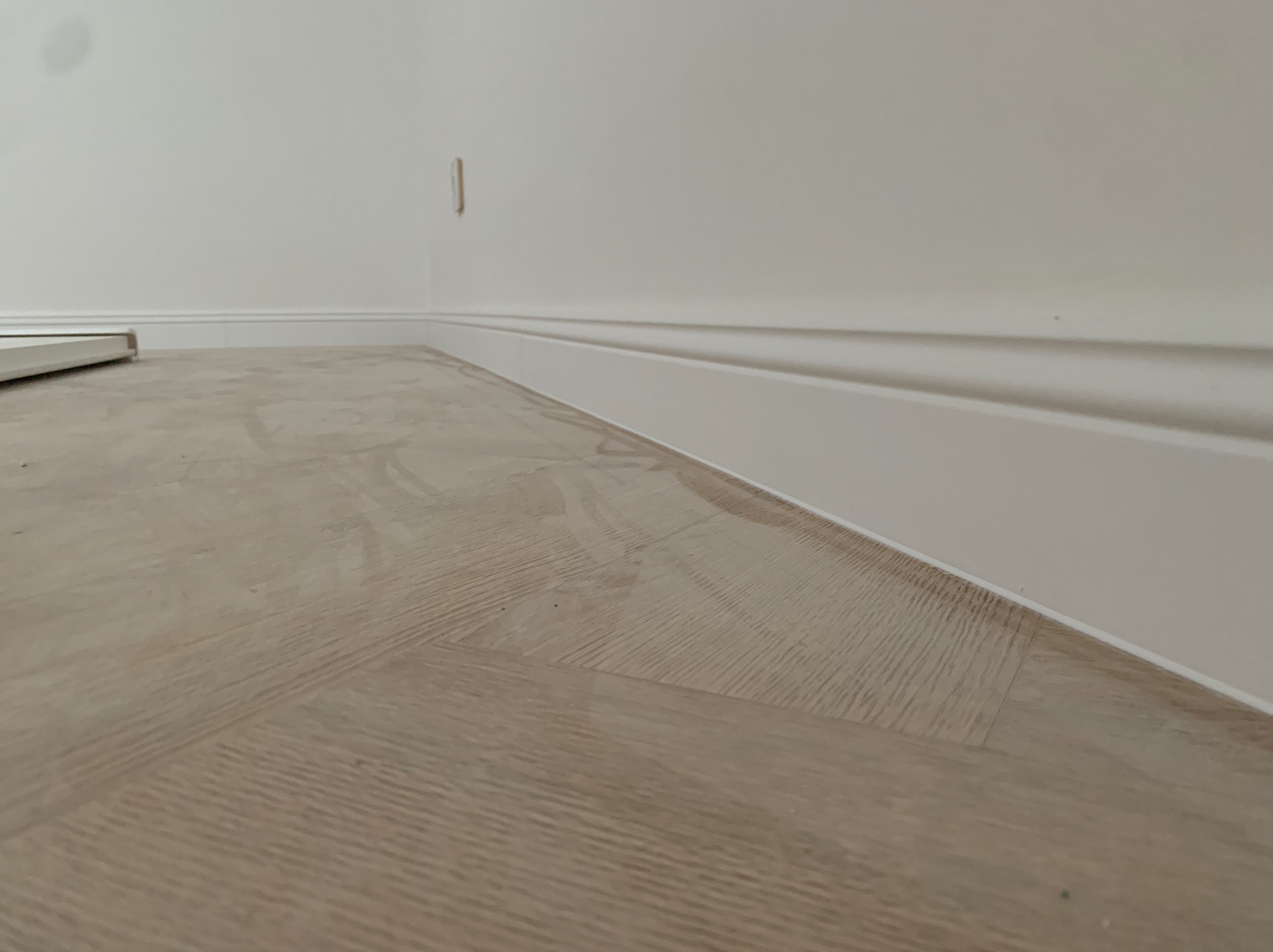 Jasna podłoga ułożona w jodełkę, białe listwy przypodłogowe przy ścianie. Widok z niskiej perspektywy, ukazujący detale wykończenia podłogi i listew.