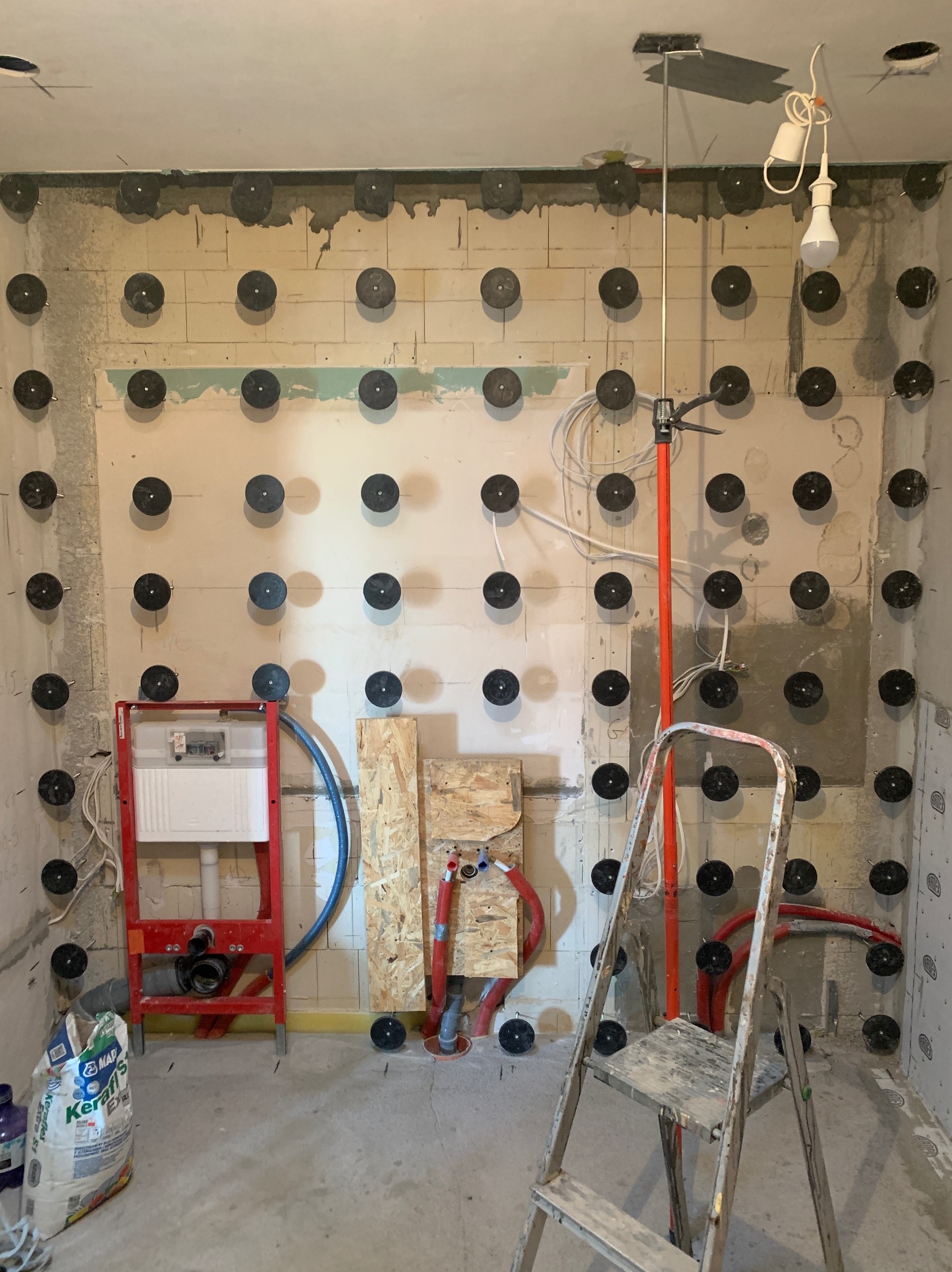 Przygotowanie ścian w łazience do położenia płytek. Widoczne mocowania, instalacja podtynkowa WC, drabina i worki z zaprawą. Ściany surowe, w trakcie prac.