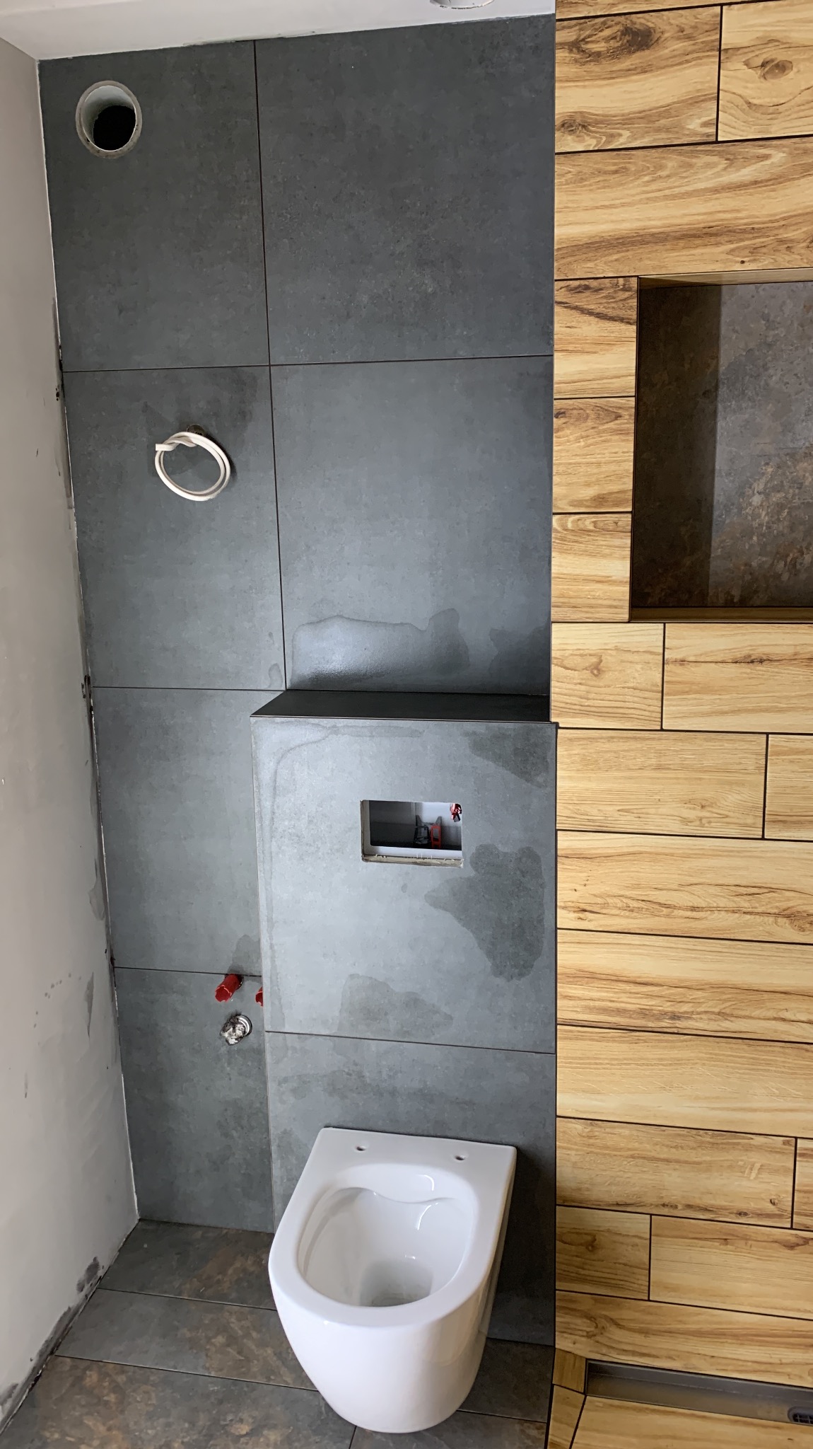 Nowoczesna łazienka w trakcie wykańczania: szare, matowe płytki na ścianie, drewnopodobne panele, podwieszana toaleta. Widoczne instalacje.