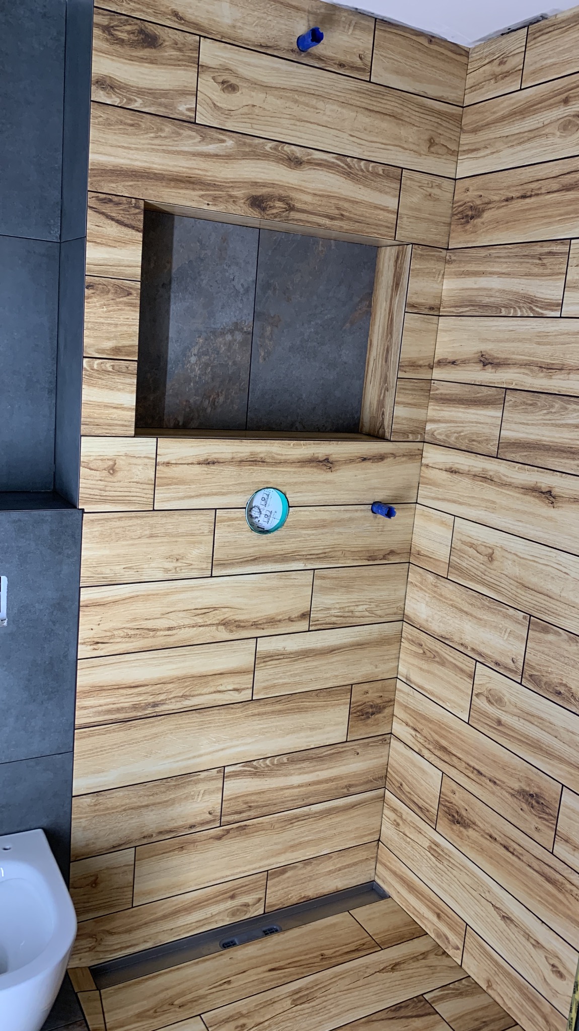 Wykończona łazienka z drewnopodobnymi płytkami, wnęką w ścianie wyłożoną szarymi płytkami i odpływem liniowym. Widoczny fragment toalety.