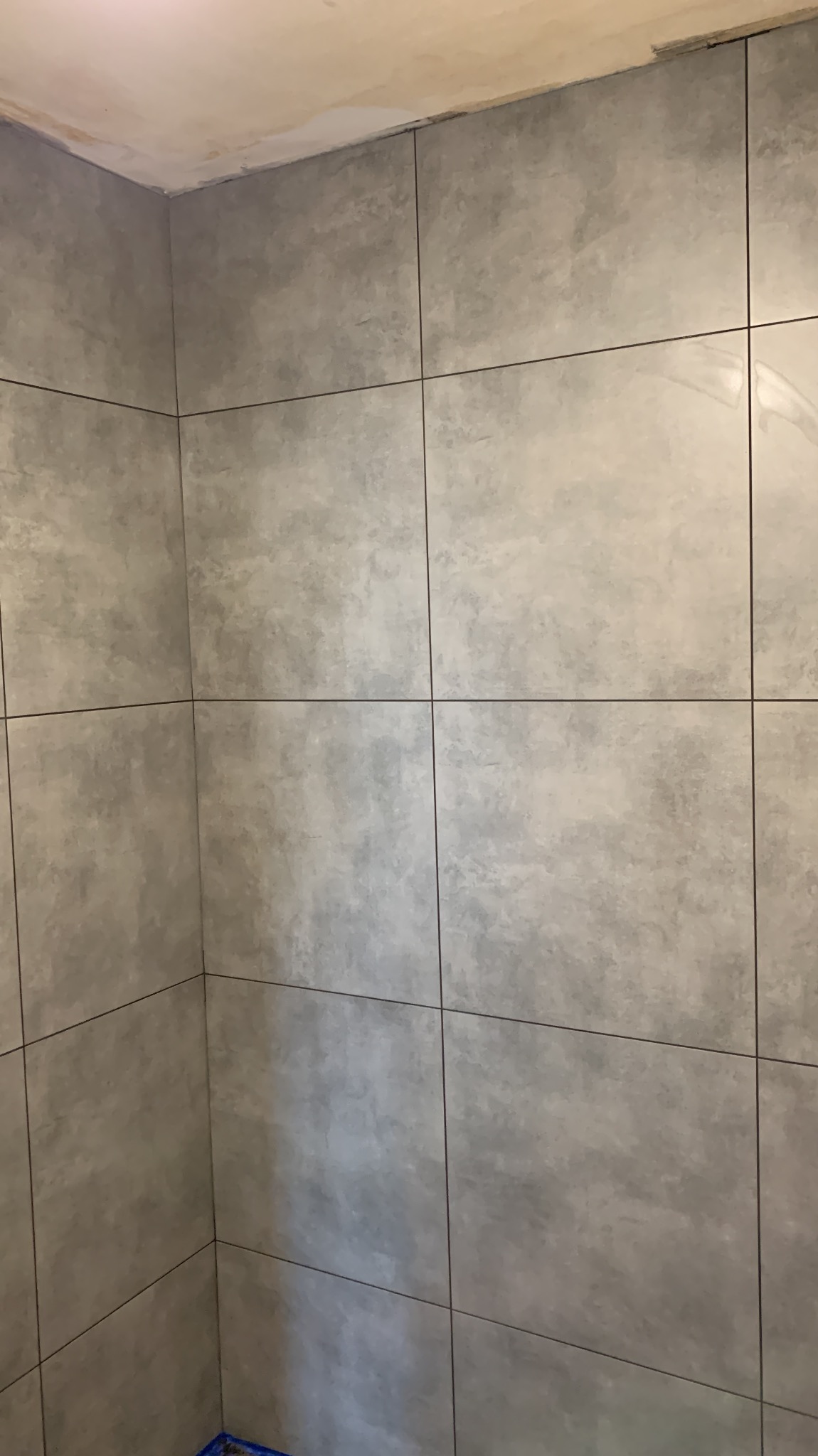 Ściana wyłożona szarymi, matowymi płytkami w łazience. Widoczny fragment sufitu przed wykończeniem. Ciemna fuga podkreśla regularny układ płytek.