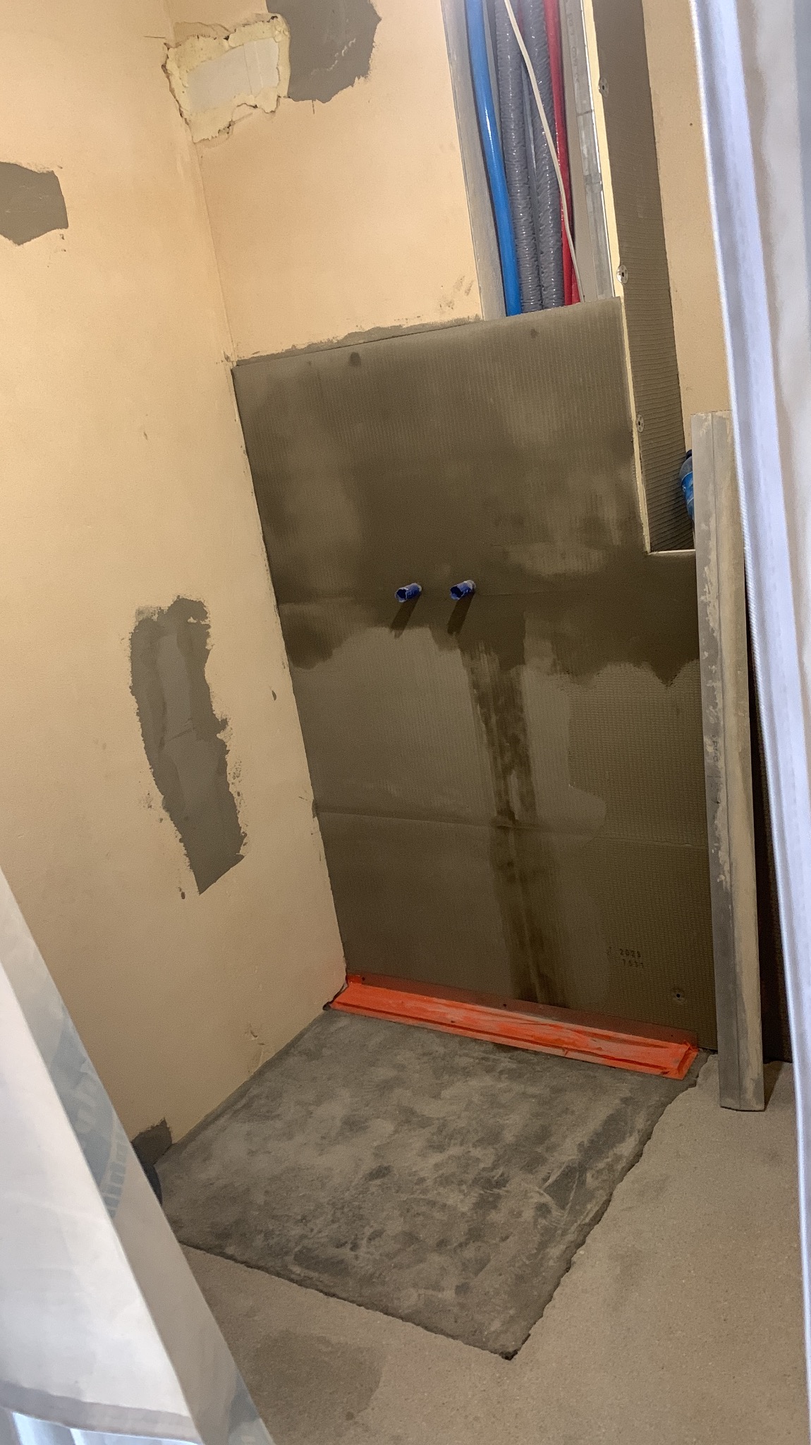 Przygotowana ściana z wyprowadzonymi rurami i wylewką pod prysznic. Widoczne ślady po szpachlowaniu na ścianie. Pomieszczenie w trakcie prac wykończeniowych.