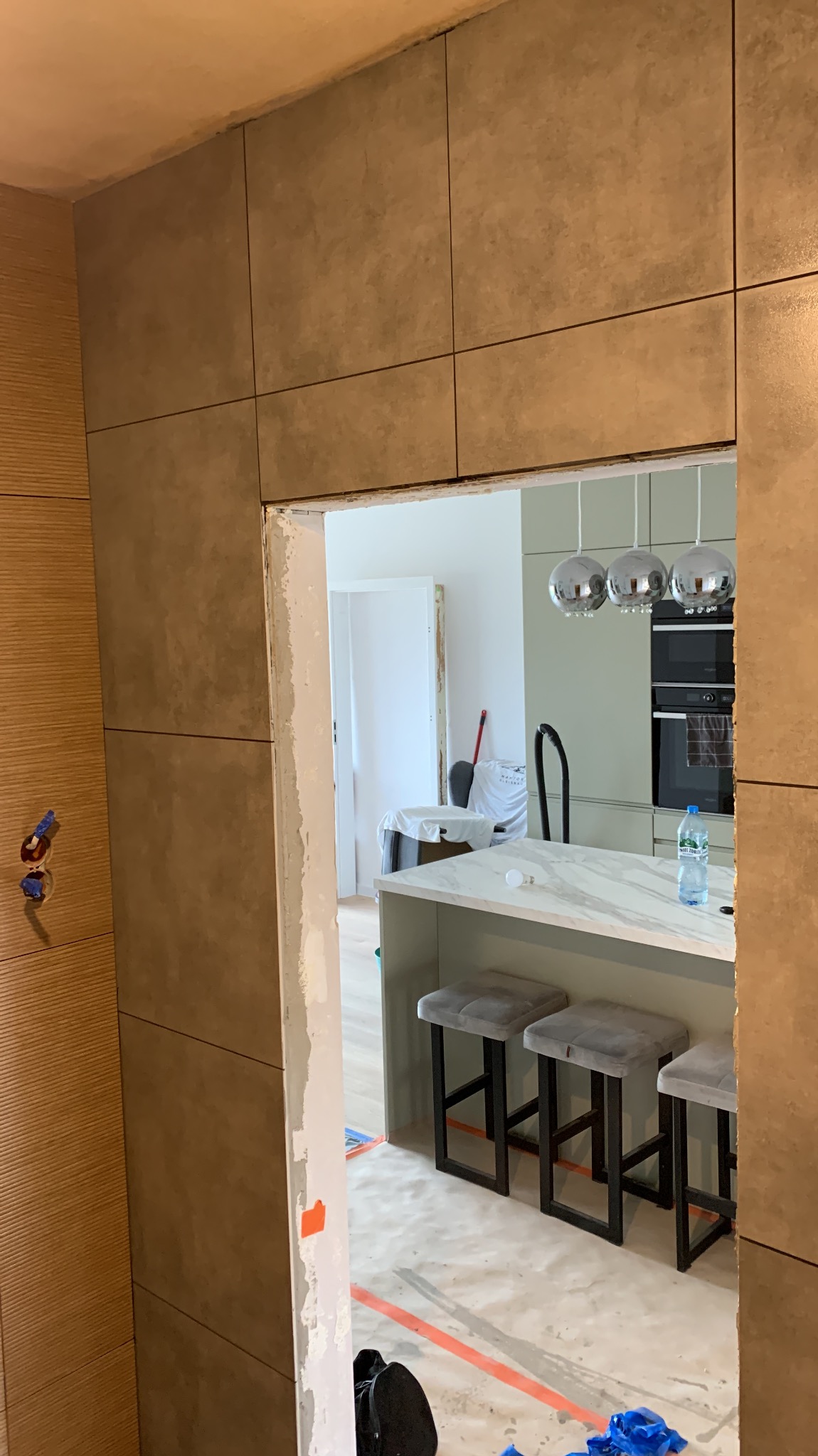 Wykończona ściana z płytek w odcieniach beżu, widok na wnętrze kuchni z wyspą i hokerami. Fragment niedokończonego przejścia z widocznymi śladami po szpachlowaniu.