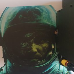 DARIUSZ NAWRAT COMMUNICATIONS - Wydruk wielkoformatowy w Bielsko-Białej: zbliżenie na twarz astronauty w hełmie, z widoczną Ziemią w odbiciu. Nietypowe ujęcie, ciemna tonacja, obok fragment maszyny drukarskiej.