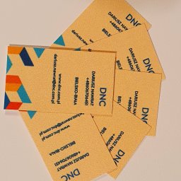 DARIUSZ NAWRAT COMMUNICATIONS - Stos wizytówek z beżowego papieru z niebieskim tekstem i geometrycznym wzorem, ułożonych pod kątem na jasnym tle. Widoczne dane kontaktowe i logo firmy DNC.