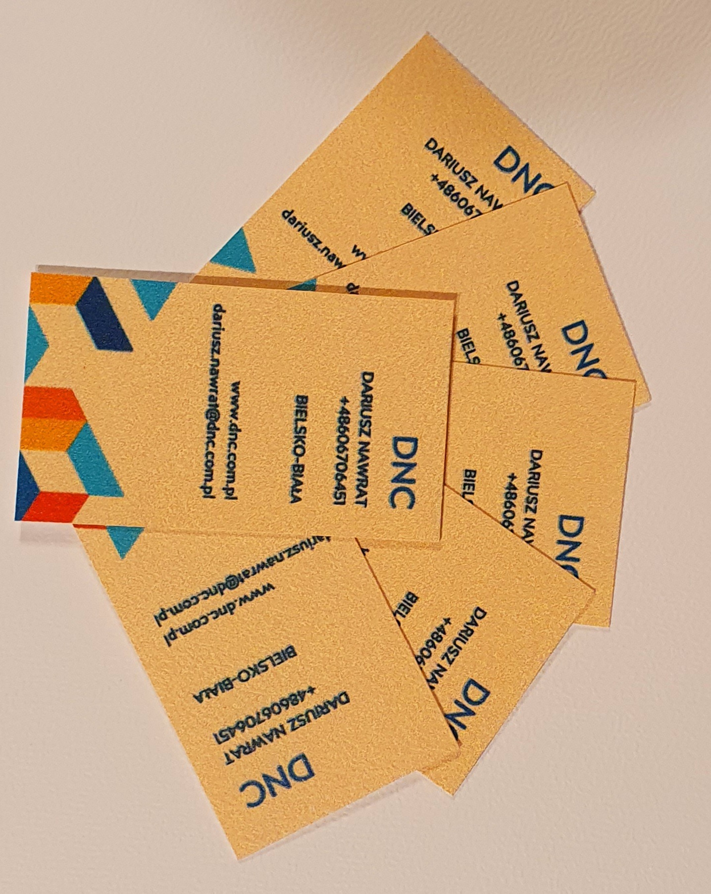 Stos wizytówek z beżowego papieru z niebieskim tekstem i geometrycznym wzorem, ułożonych pod kątem na jasnym tle. Widoczne dane kontaktowe i logo firmy DNC.