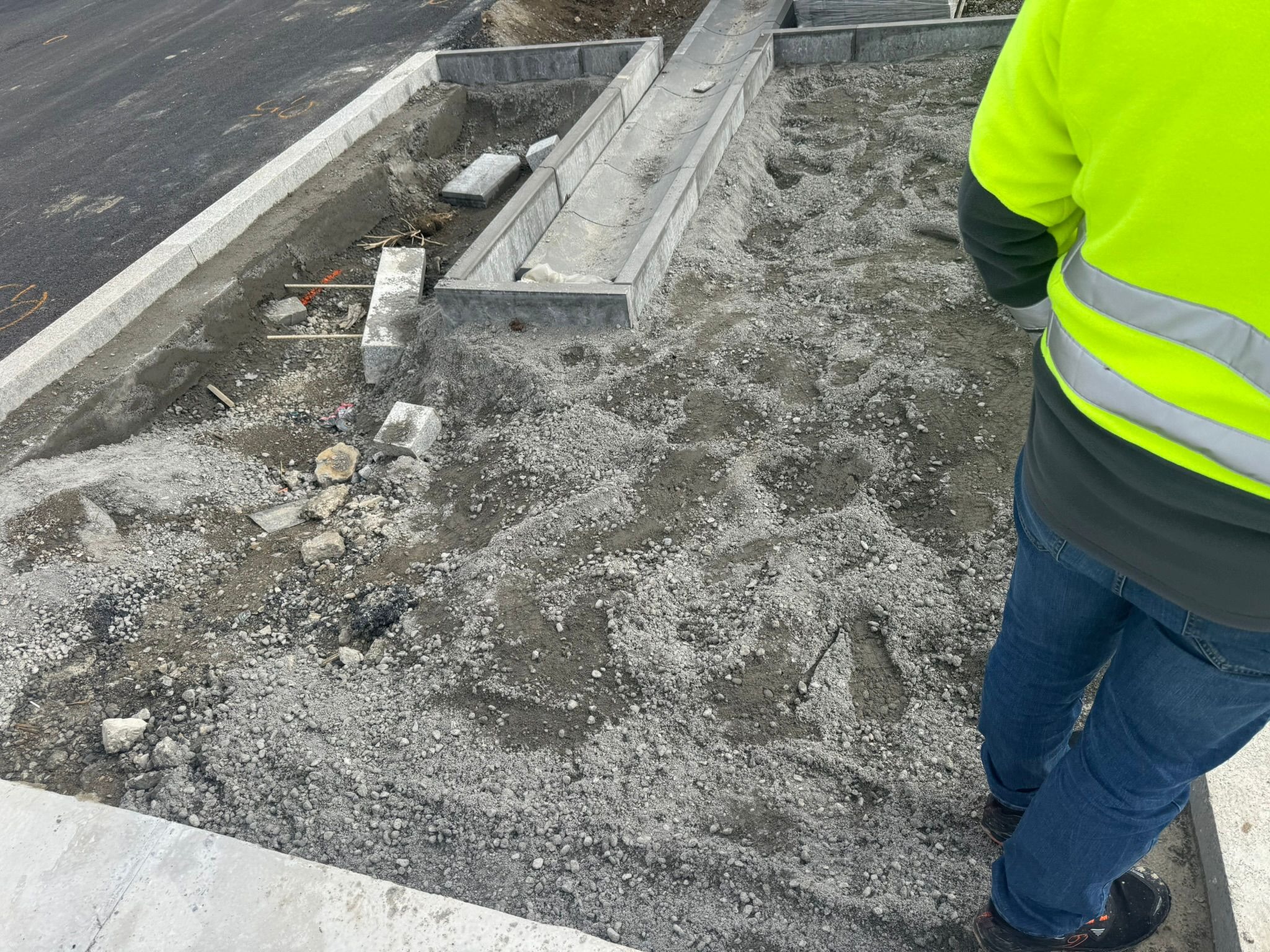 Przygotowanie podłoża pod kostkę granitową z ułożonym korytkiem odwadniającym i obrzeżami. Widoczny pracownik w odblaskowej kamizelce.