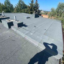 REVENTO - Szary dach pokryty papą termozgrzewalną z kominami wentylacyjnymi i brązową rynną. Cień osoby w prawym dolnym rogu. Widok z góry na budynek i zieleń w tle.