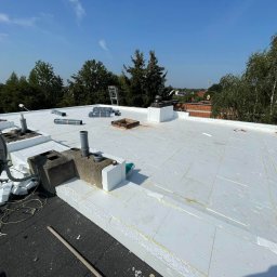 REVENTO - Białe płyty ociepleniowe na dachu z kominami i anteną satelitarną, widok z góry w słoneczny dzień. Częściowo widoczna drabina i zieleń w tle.