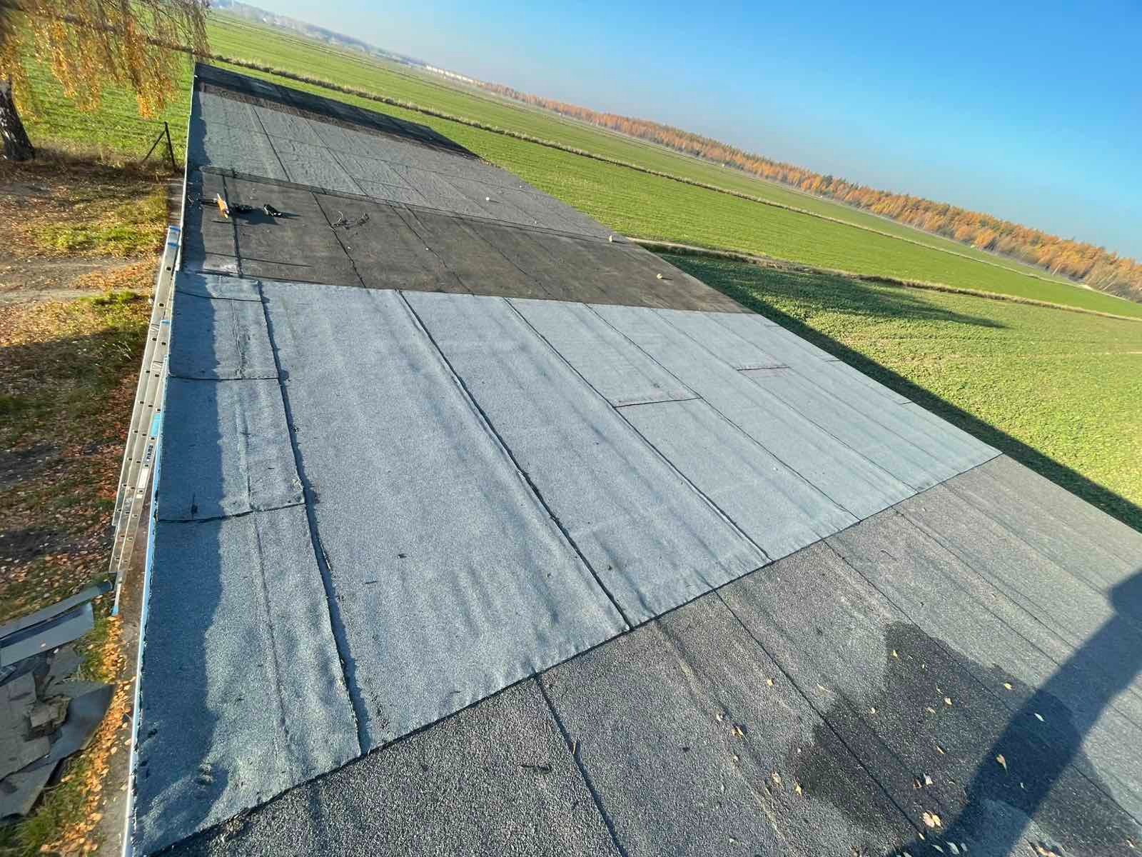 Widok z góry na fragment dachu pokrytego papą w różnych odcieniach szarości, z widoczną drabiną i polami w tle. Część dachu jest świeżo położona.