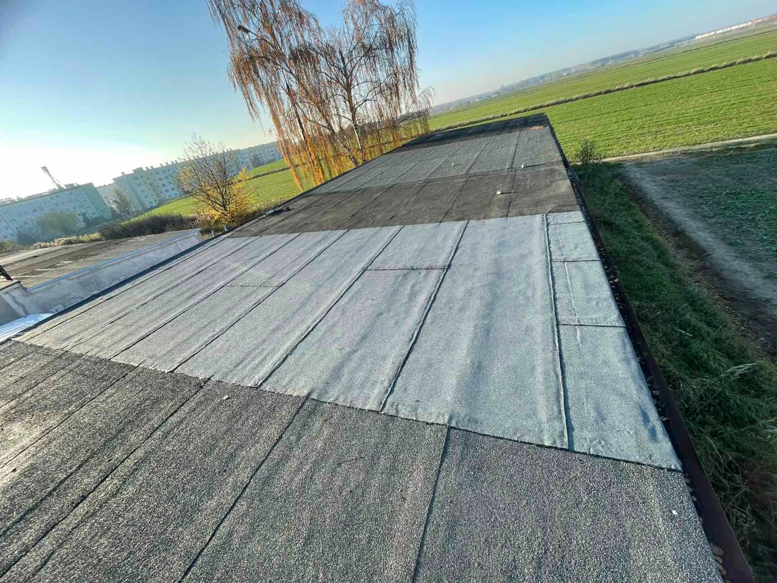 Wykończony dach płaski pokryty papą w różnych odcieniach szarości, z widocznym tłem zielonych pól i budynków mieszkalnych w oddali, w słoneczny dzień.